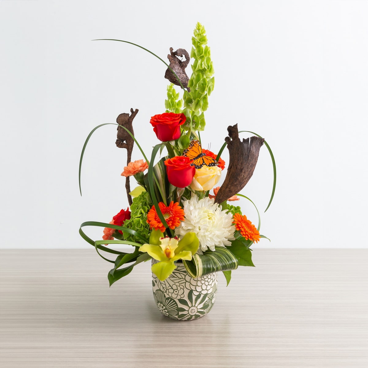 Nature’s Symphony floral arrangement featuring red and orange roses, green hydrangea, bells of Ireland, tropical accents, and decorative wood elements in a decorative vase, with dimensions of 17 inches high by 10 inches wide."