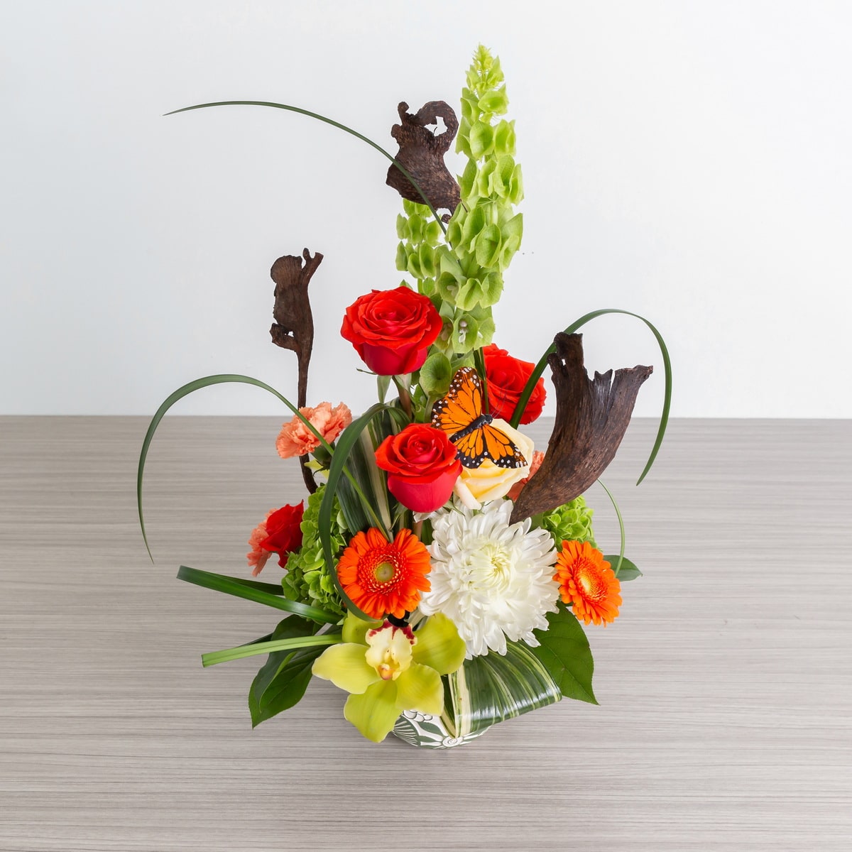 Nature’s Symphony floral arrangement featuring red and orange roses, green hydrangea, bells of Ireland, tropical accents, and decorative wood elements in a decorative vase, with dimensions of 17 inches high by 10 inches wide."