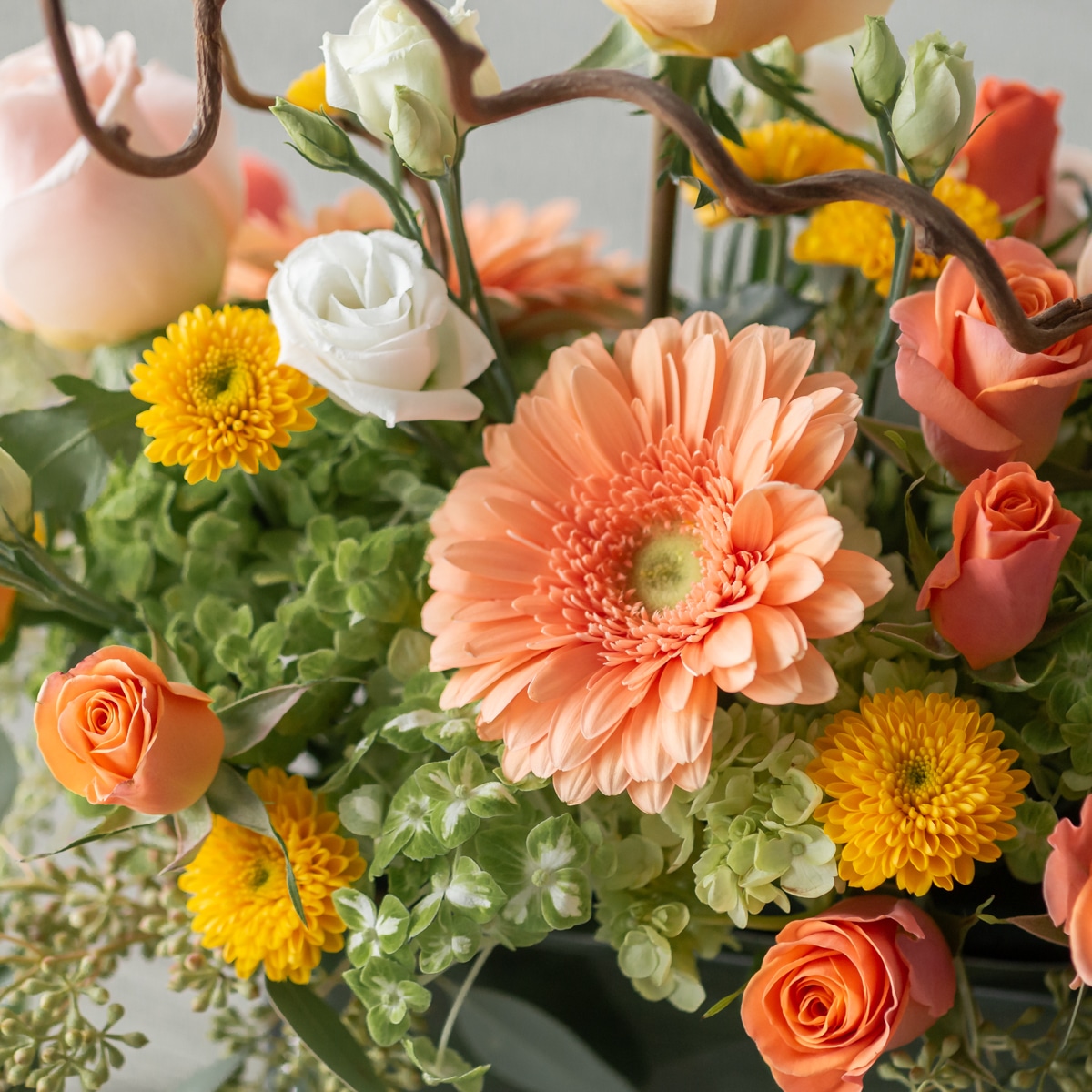 Graceful Elegance floral arrangement with peach roses, gerbera, white lisianthus, yellow accents, and wooden branches in a rectangular vase