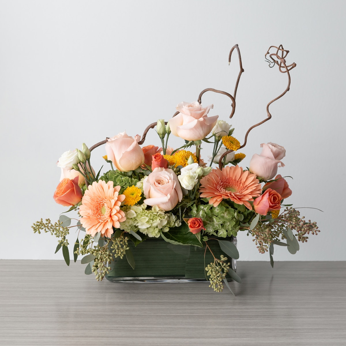 Graceful Elegance floral arrangement with peach roses, gerbera, white lisianthus, yellow accents, and wooden branches in a rectangular vase