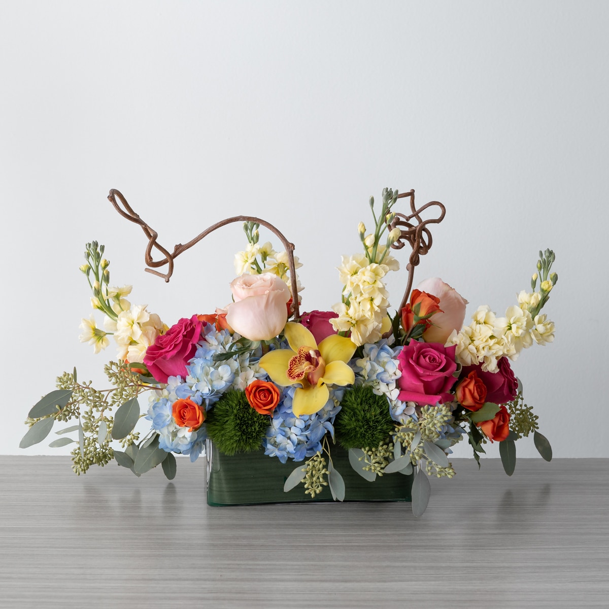 Charming Delight floral arrangement featuring pink and orange roses, yellow orchids, hydrangeas, stock flowers, and decorative wooden branches, measuring 15 inches long, 12 inches high, and 9 inches wide