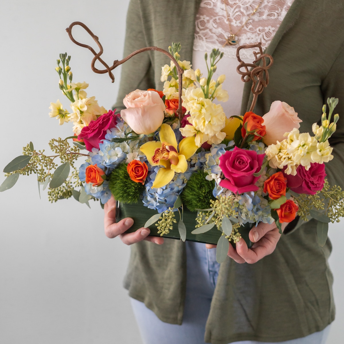 Charming Delight floral arrangement featuring pink and orange roses, yellow orchids, hydrangeas, stock flowers, and decorative wooden branches, measuring 15 inches long, 12 inches high, and 9 inches wide.